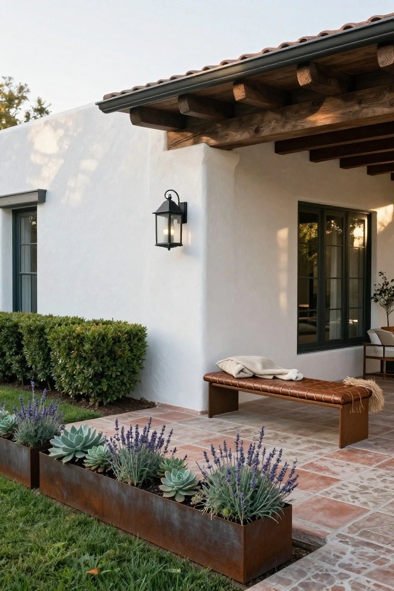 White stucco wall of a house with dark green windows and a covered porch featuring a brown leather bench with white throw, black lantern light, boxwood shrubs, and linear rusted metal planters filled with succulents and lavender on terracotta tile patio.
