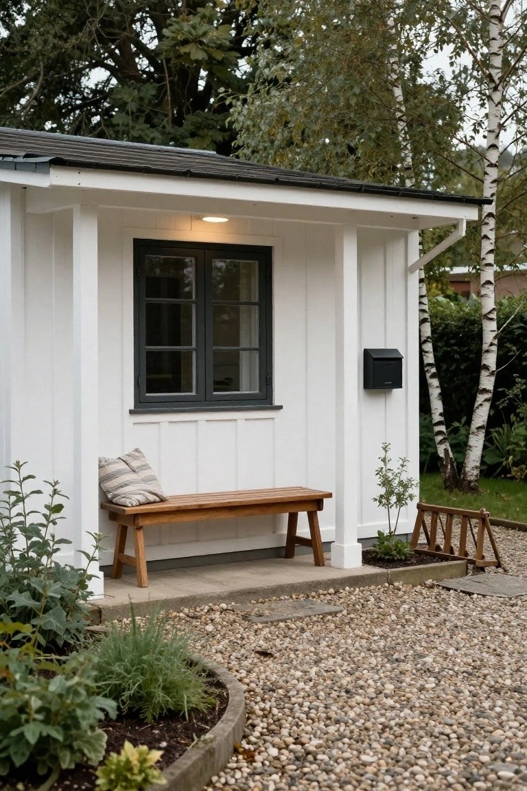 White clapboard cottage exterior with covered porch, black-framed window, wooden bench with striped cushion, black mailbox, plants, and gravel path.