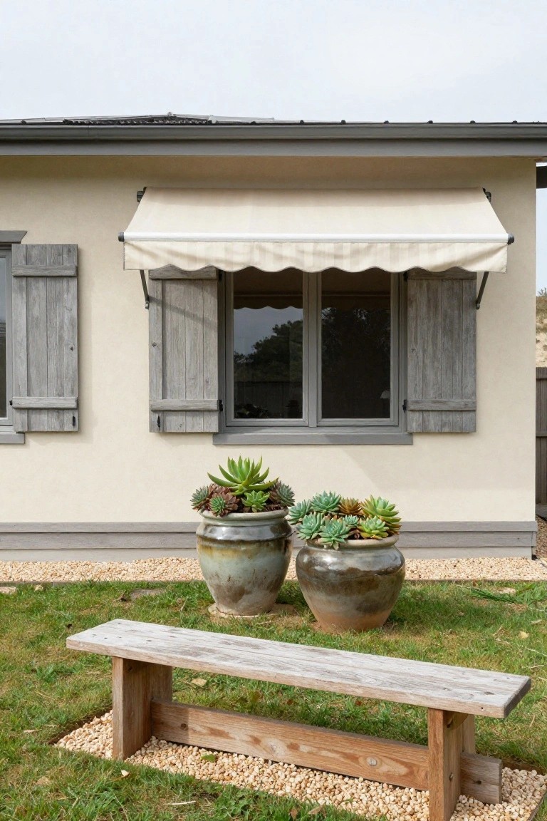 Beige stucco house wall with gray metal roof, wooden shutters framing a window with beige awning, two large terracotta pots filled with succulents, and a weathered wooden bench on grass surrounded by gravel.