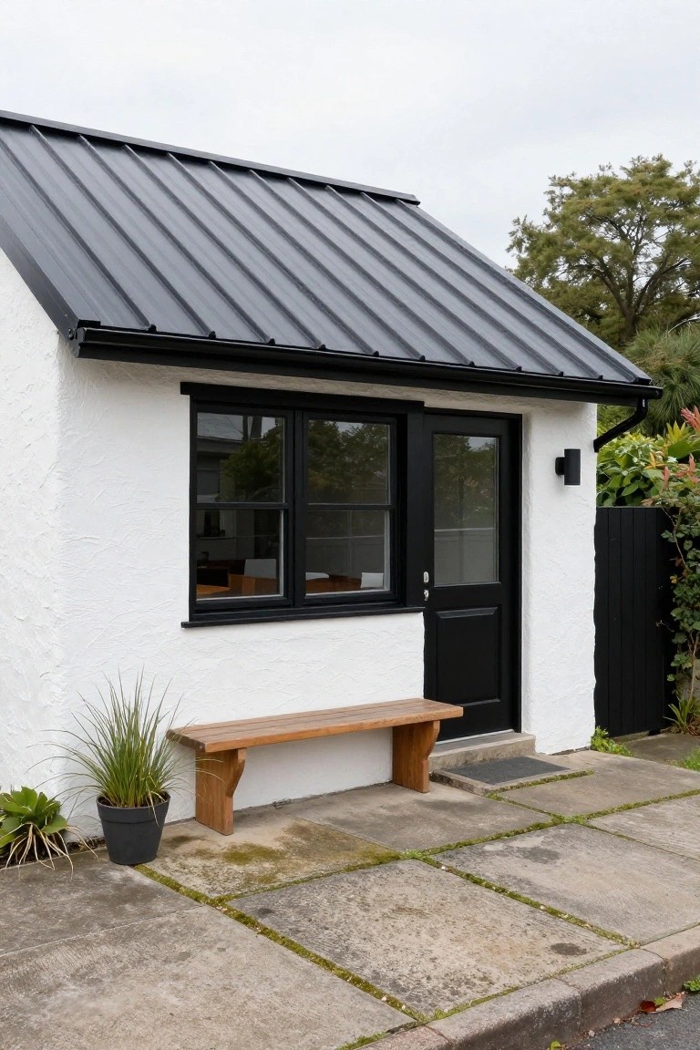 White stucco-walled house with black corrugated metal sloped roof, black-framed double window and door, wooden bench, potted grass plant, and concrete paver path.
