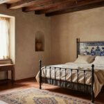 Bedroom interior with exposed dark wooden ceiling beams, white plaster walls, arched window with sheer curtains, iron bed with colorful tiled headboard and white linens, wooden furniture, arched niche holding terracotta vase, Persian rug on terracotta tile floor.