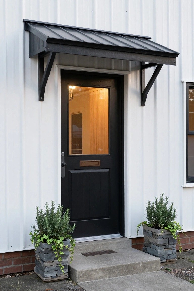 White metal-sided house exterior featuring a black metal awning over a black front door with glass panel, flanked by two potted rosemary plants in stacked stone planters on concrete steps.