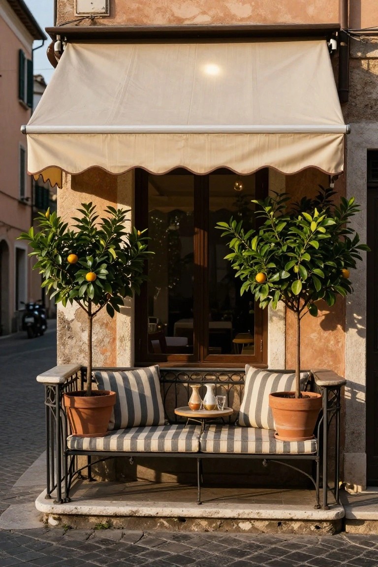 Wrought iron bench with beige and white striped cushions on a balcony, flanked by two potted orange trees in terracotta pots, small round table with glasses and vases nearby, under beige awning in front of glass doors on beige stucco building.