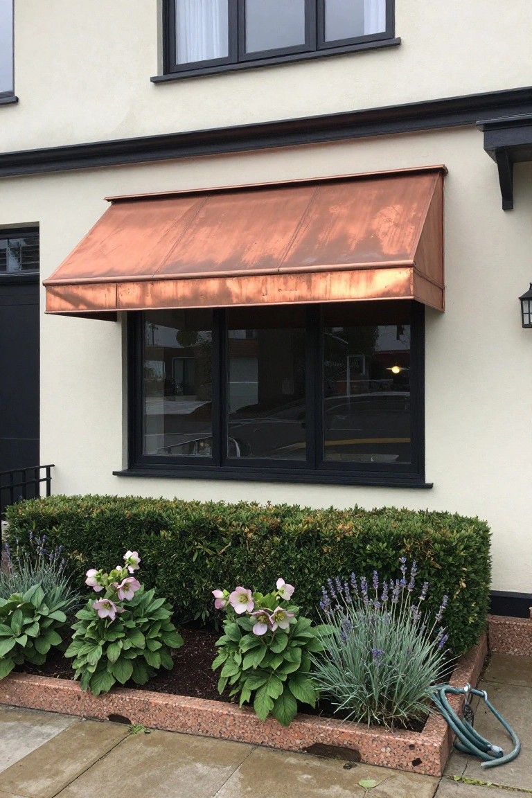 House exterior with a patinaed copper awning extending over a black-framed window on white walls, black trim above, and a landscaped bed of boxwood hedges, hosta plants, and lavender below.