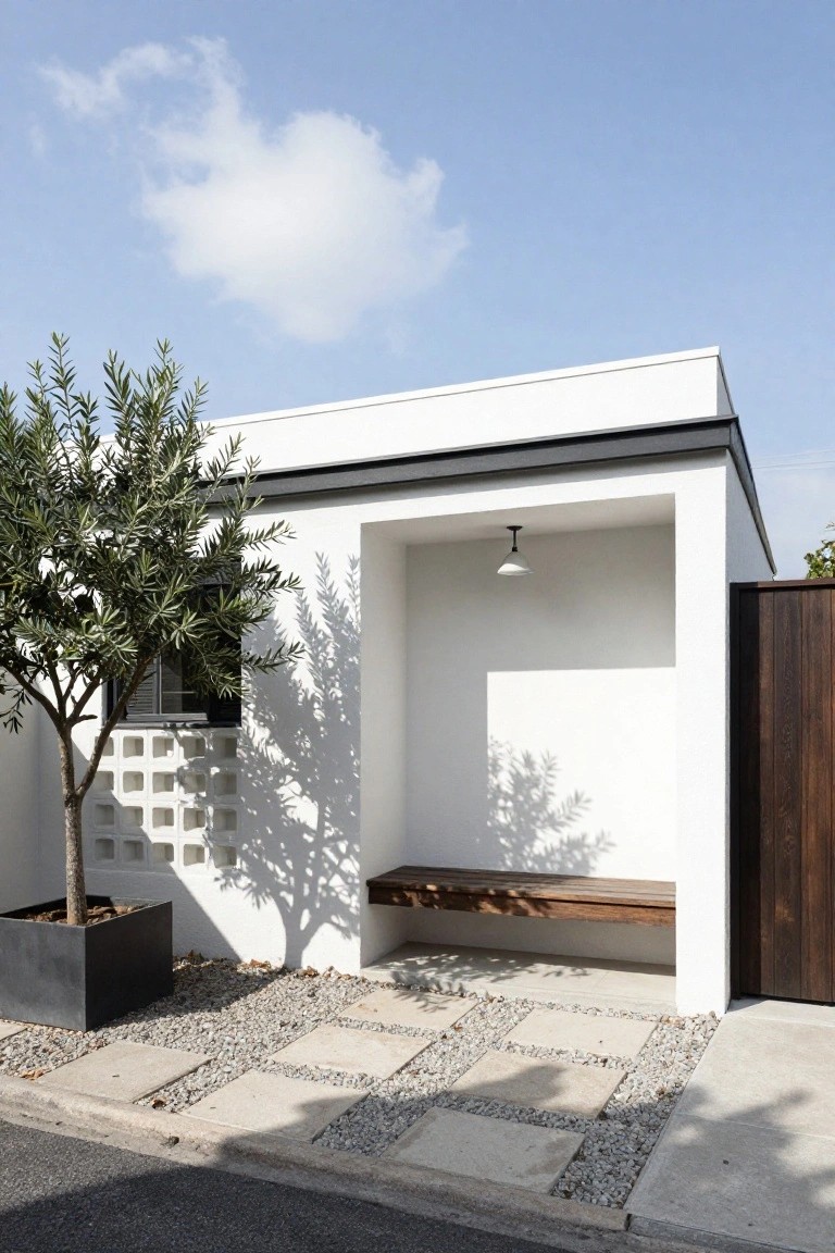 White stucco house exterior with recessed open alcove containing wooden bench and hanging light, potted olive tree casting shadows on block window and walls, gravel and pavers on ground leading to wooden fence gate.