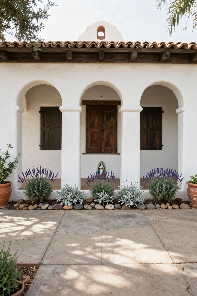 White stucco Spanish Colonial house facade with terracotta tile roof, three arched porch openings, central dark wooden double doors, side wooden shutters, Virgin Mary statue niche above door, lavender plants in terracotta pots, and gravel border on concrete pavers.