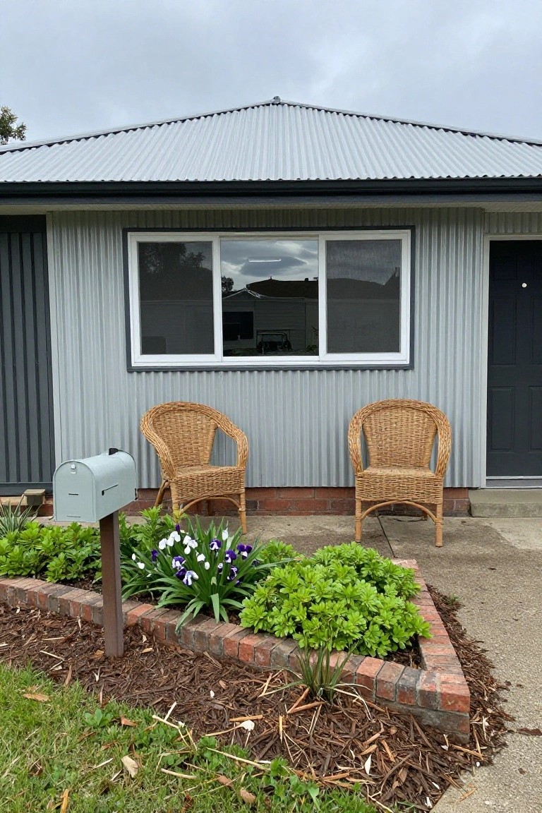 Gray corrugated metal house exterior with sloped roof, large white-framed window, black front door, two wicker armchairs on concrete porch, blue mailbox on post, and brick-edged garden bed with green plants and purple flowers.