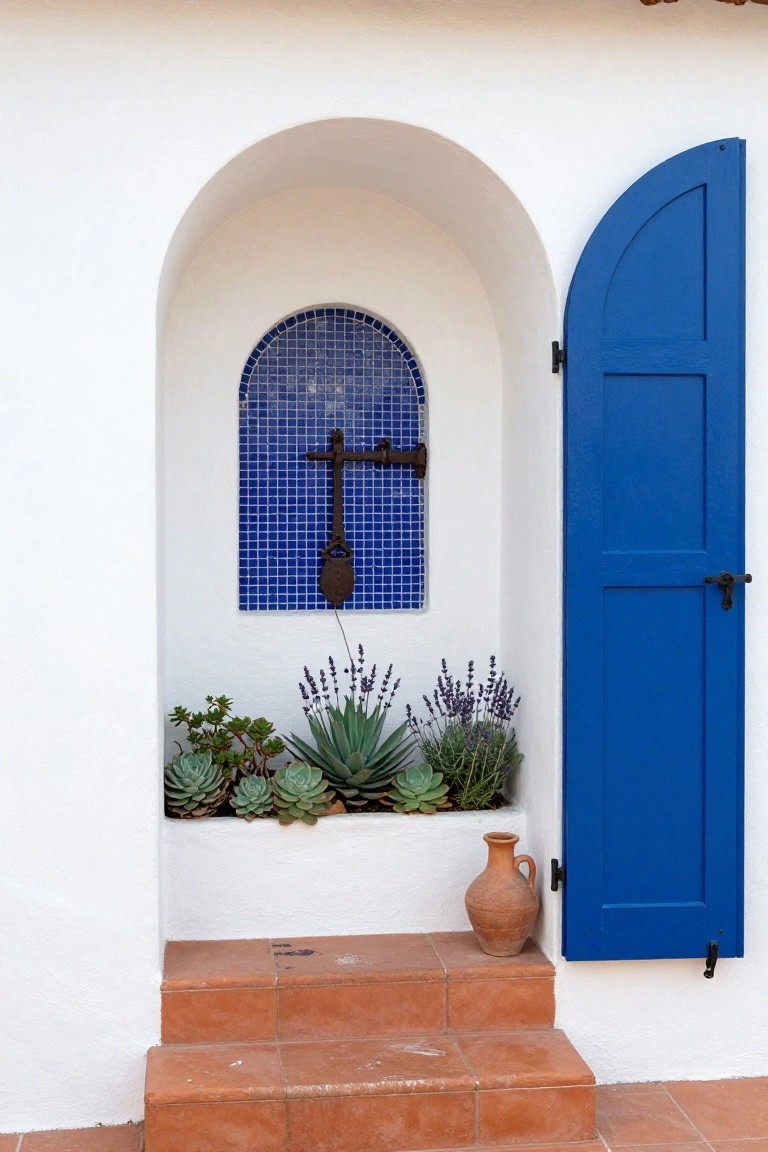 White stucco arched doorway with blue mosaic tiled niche containing iron cross, flanked by open blue shutters, lavender and succulent plants in planter box, terracotta pot, and steps.
