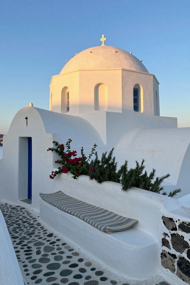 White domed building with arched entry featuring a blue door, red bougainvillea and green shrubs along the wall, a cushioned bench beside the door, and a pebble stone path leading to it under a clear blue sky.