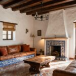 Hacienda-style living room interior with white stucco walls, exposed wooden beams, blue-and-white tiled fireplace, patterned terracotta tile floor, brown leather sofas, wooden coffee table, potted olive tree, and arched window with bamboo shade.