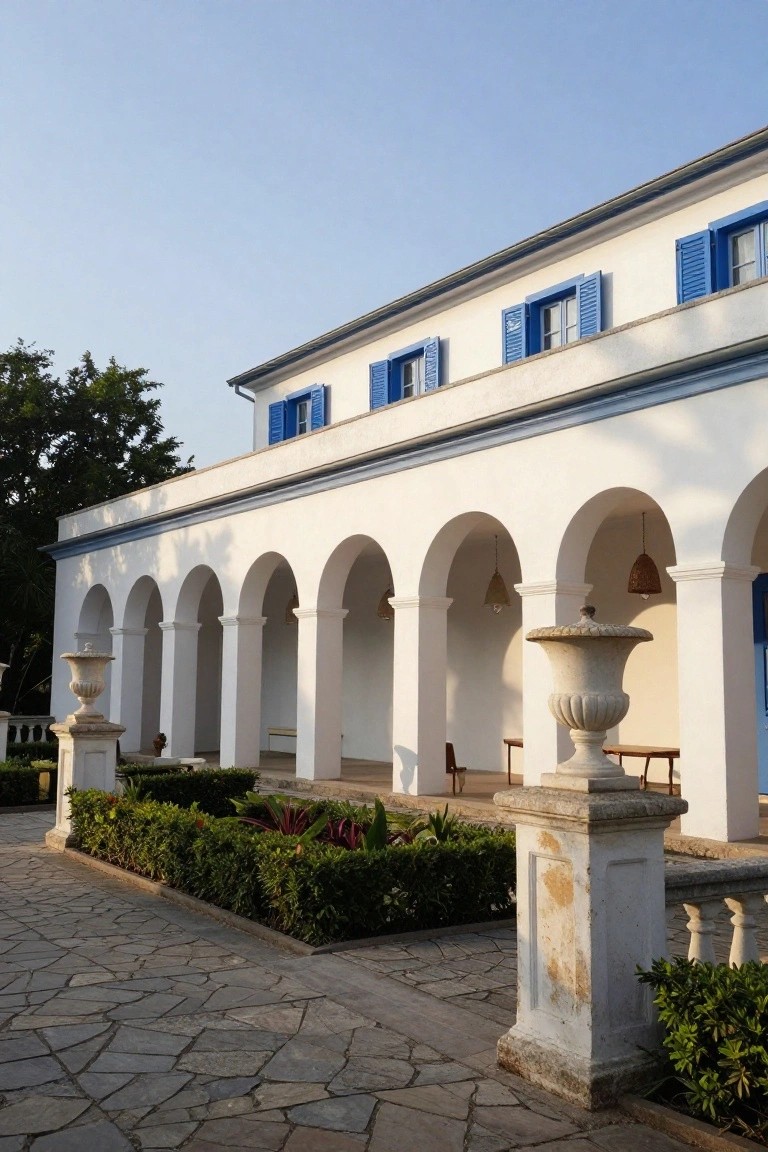 White stucco house with blue shutters and a row of arched colonnade supported by columns, flanked by stone urns and low hedges on a stone-paved walkway.