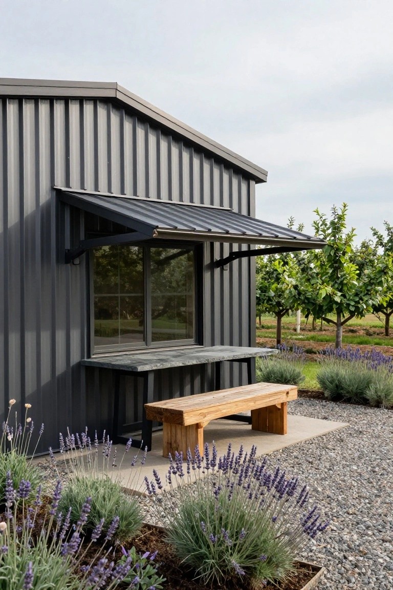 Gray corrugated metal building side with black awning over a window featuring a stone countertop extension, wooden bench in front, lavender plants in gravel beds, and orchard trees in the background.