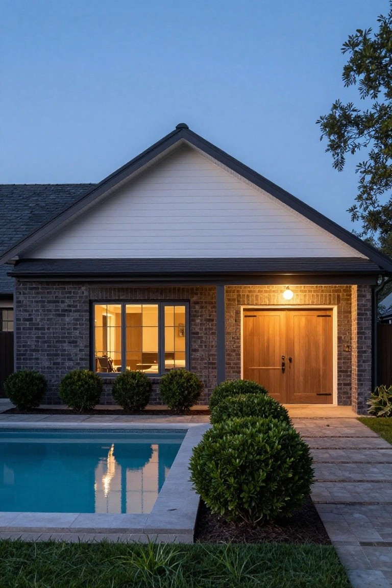 Evening photo of a ranch-style house with dark brick lower walls, white horizontal siding on the gabled roofline, double wooden entry doors under a lit porch, boxwood shrubs, a turquoise pool, and paver pathway.