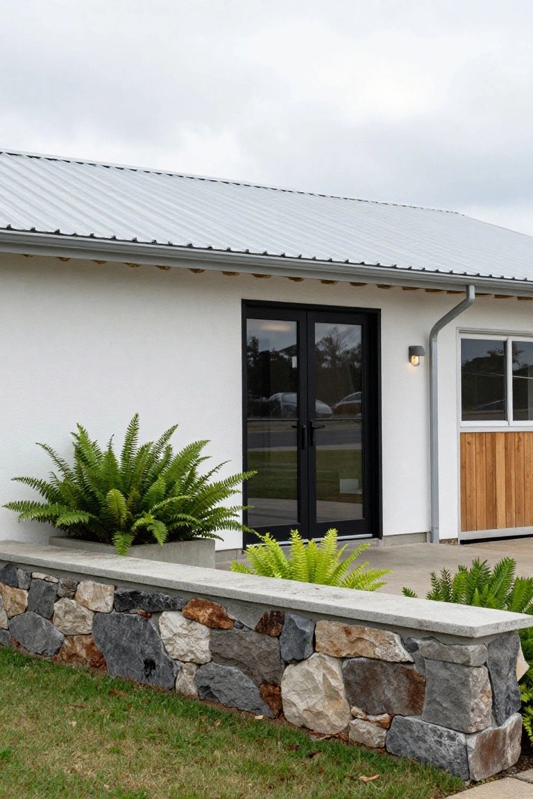 White stucco ranch house exterior with black-framed glass double doors, low stone retaining wall topped with concrete and planted with ferns, metal roof overhang, adjacent wood-paneled door, and grass lawn.