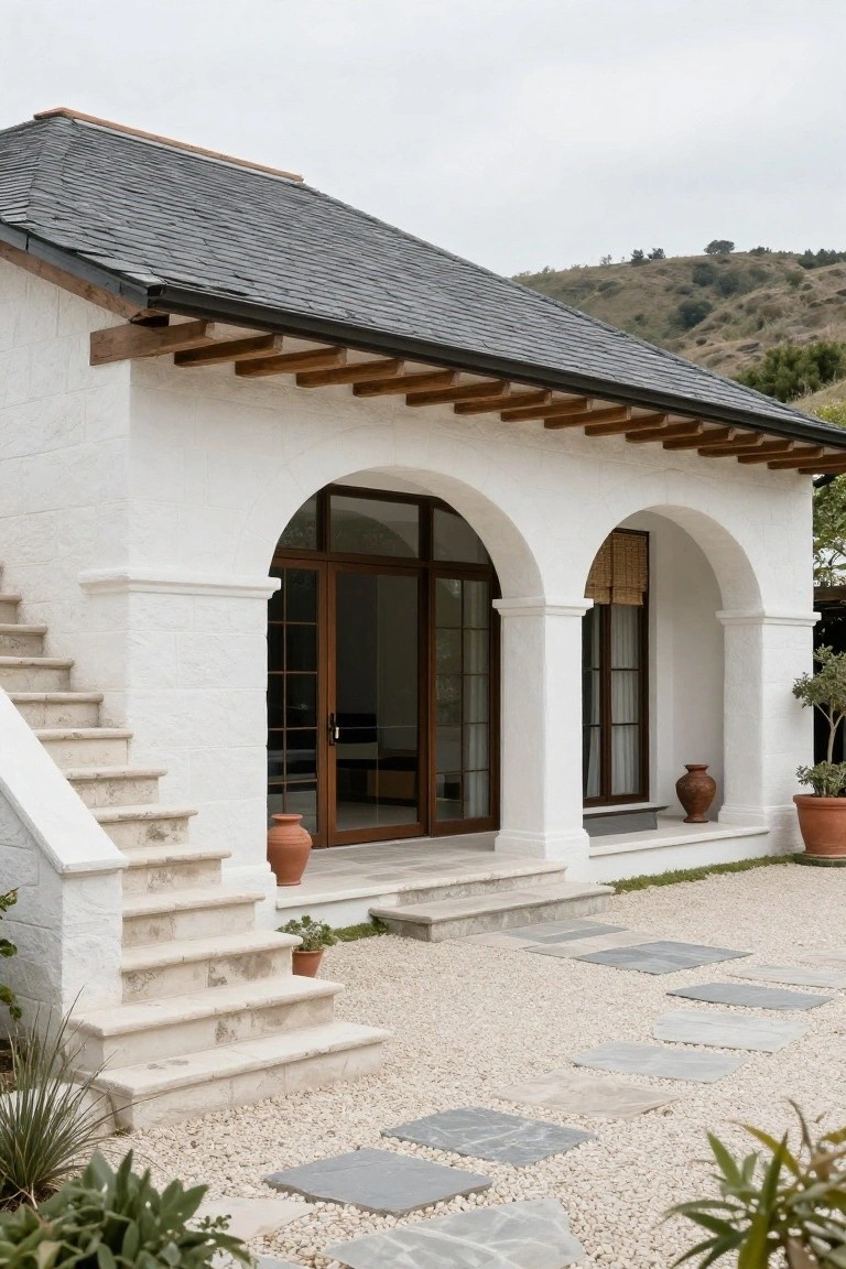 White stucco house exterior featuring double arched doorways with wooden doors and windows, flanked by columns, stone steps, gravel ground with stone pavers, potted plants, and a hillside in the background.