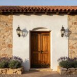 Hacienda-style house exterior featuring a wooden double door entry with iron hardware, flanked by lanterns on stucco walls with stone base, potted lavender plant, and stone fountain nearby.