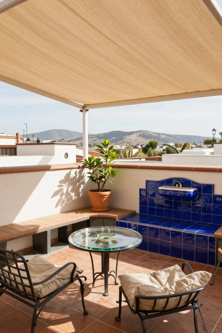 Rooftop terrace with beige shade canopy overhead, white stucco wall featuring built-in terracotta bench and blue tiled fountain, round glass table, two black wrought iron chairs with beige cushions, potted plants, and background view of hills and buildings.