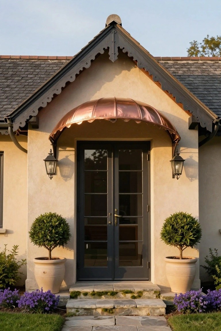 Front entrance of a beige stucco house with dark gray slate roof, curved copper awning over black double doors, lanterns on walls, potted boxwood topiaries, stone steps, and purple flower clusters in front.