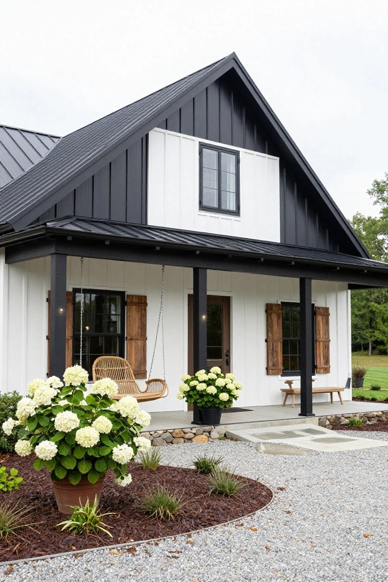 Front exterior of a modern farmhouse with black metal gable roof and siding accents, white board-and-batten walls, covered porch featuring a hanging swing and bench, flanked by potted white hydrangeas on a gravel driveway with mulch beds.
