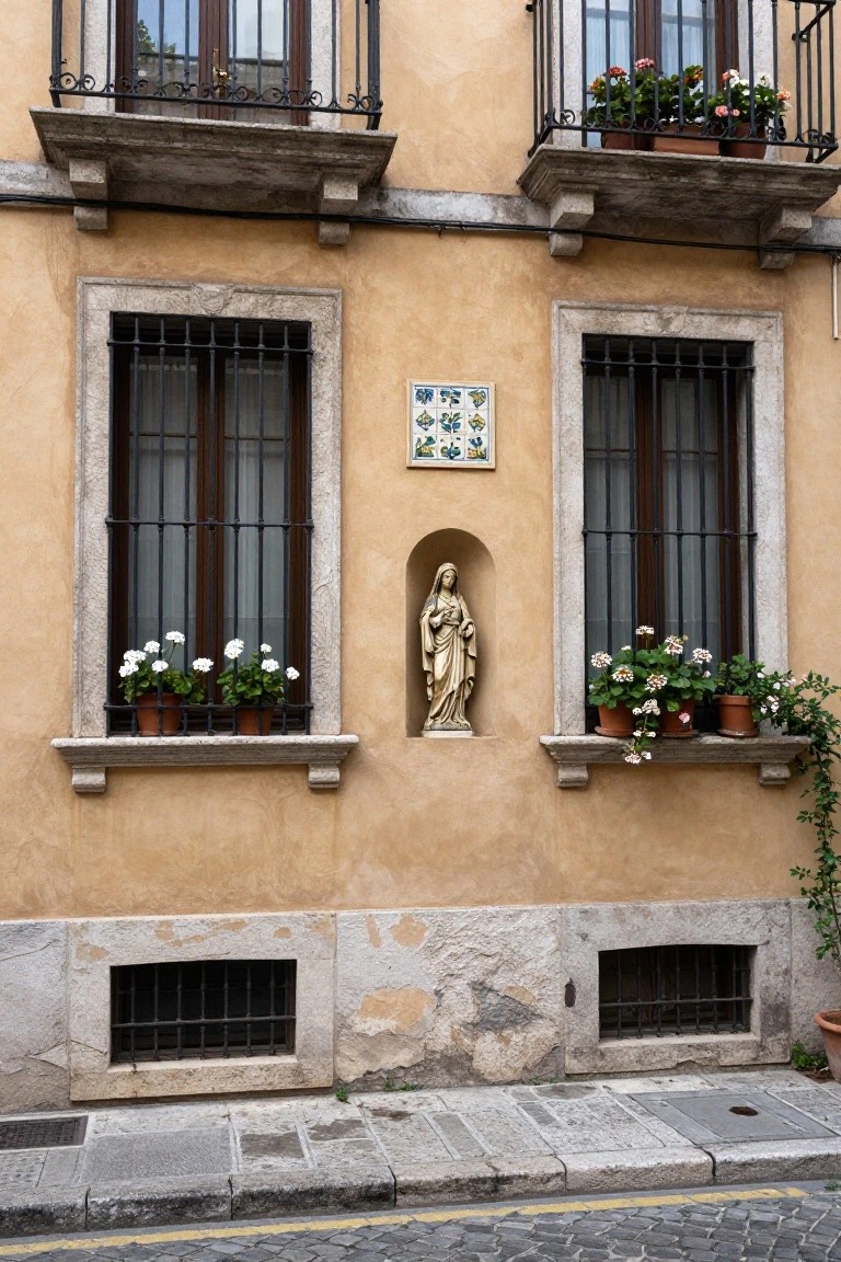 Ochre stucco building facade with wrought-iron window grilles and balconies overflowing with potted flowers, a central niche containing a golden Virgin Mary statue, a blue tiled plaque, and lower basement grilles, set against a cobblestone street.