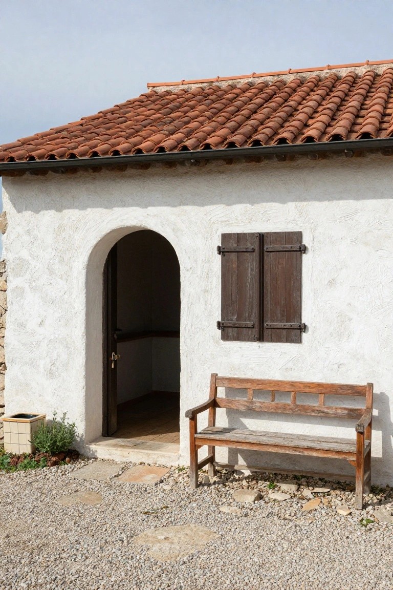 White stucco building with arched doorway, wooden bench, and red-tiled roof.