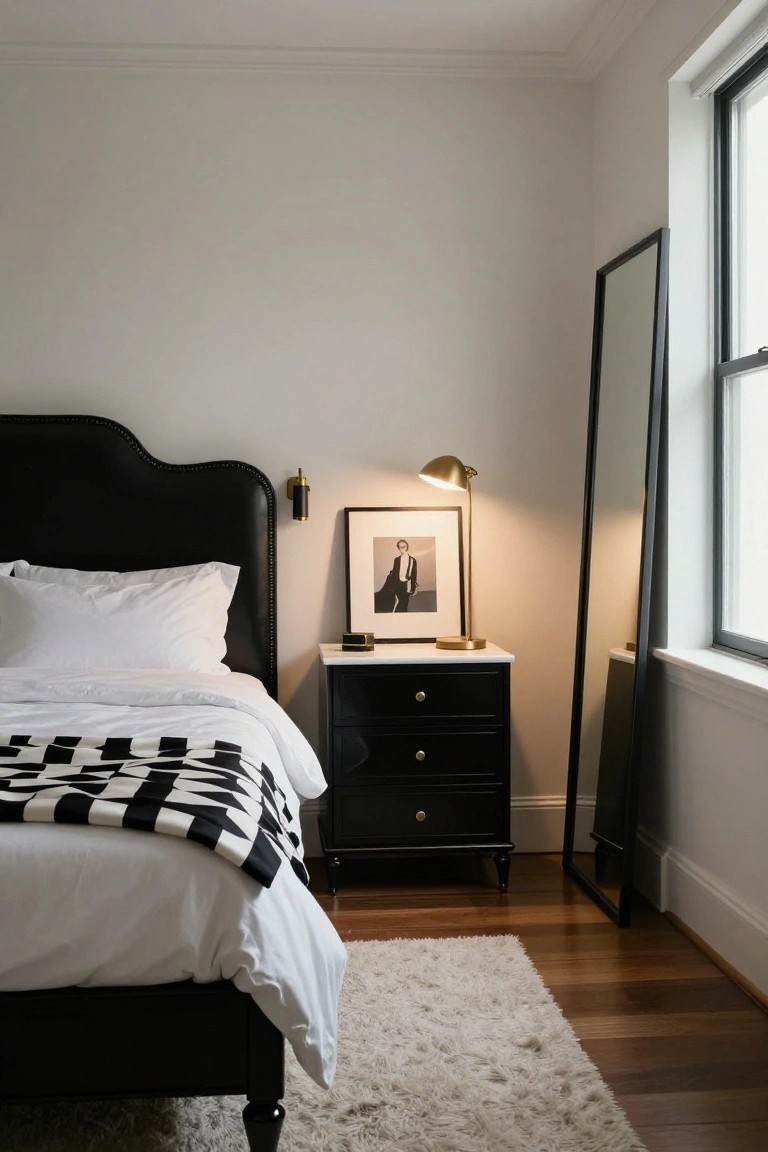 Bedroom with black tufted leather headboard on bed covered in white sheets and black-and-white checkered throw blanket, black wood nightstand holding gold lamp and framed photo of man in suit, tall black floor mirror next to window, light walls, and wood floor with cream rug.