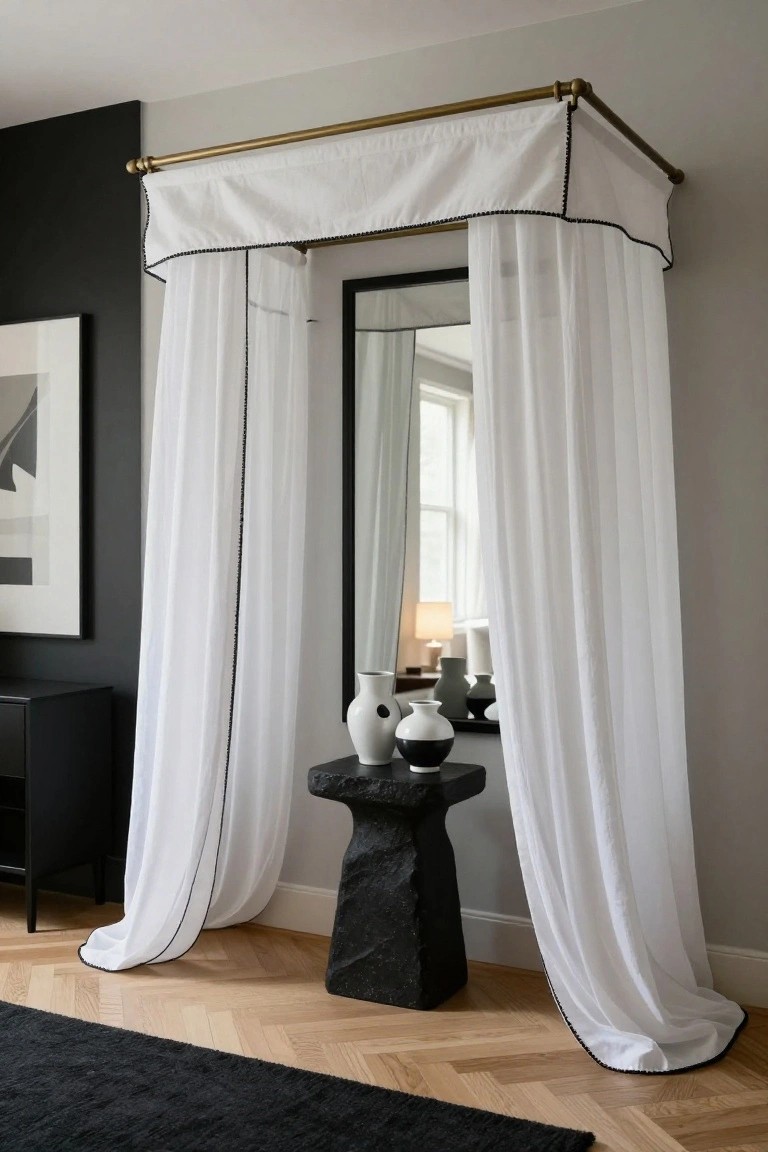 Brass pipe canopy frame with white draped curtains surrounding a full-length mirror, a black stool holding white and black vases below, against a dark wall in an interior room with herringbone wood floor.