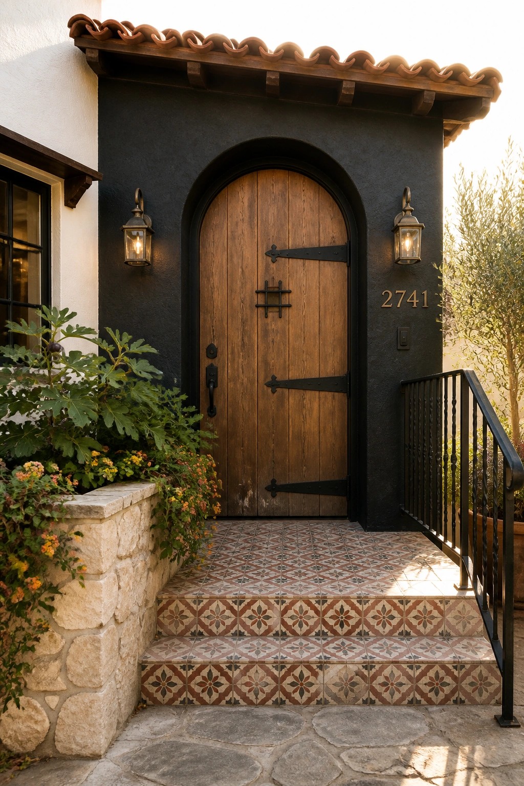 Front entry of a small home featuring an arched wooden door recessed in a dark stucco wall, with patterned tile steps and wall-mounted lanterns.