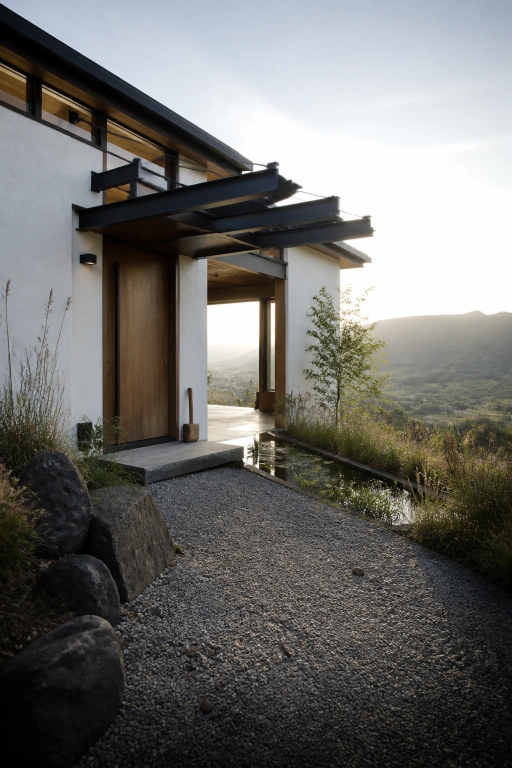 A modern white house with a wooden front door under a black metal overhang, a gravel path bordered by large rocks and native plants, and a mountain valley view in the background.