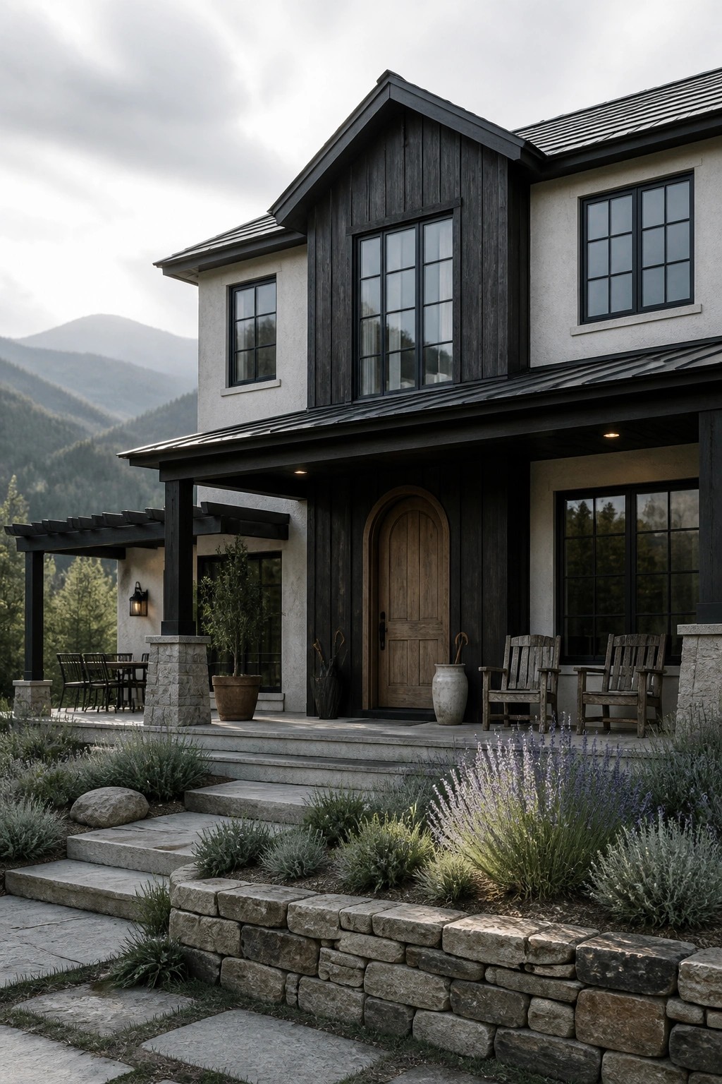 A two-story mountain home exterior with dark wood siding on the gable and entry surround, light stucco walls, a wooden arched door, covered porch with rocking chairs, and stone retaining walls with landscaping in the foreground.