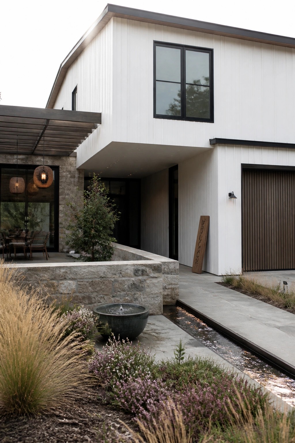 Modern white house exterior with black windows, stone retaining wall, linear water channel, and mixed native plantings along the front.