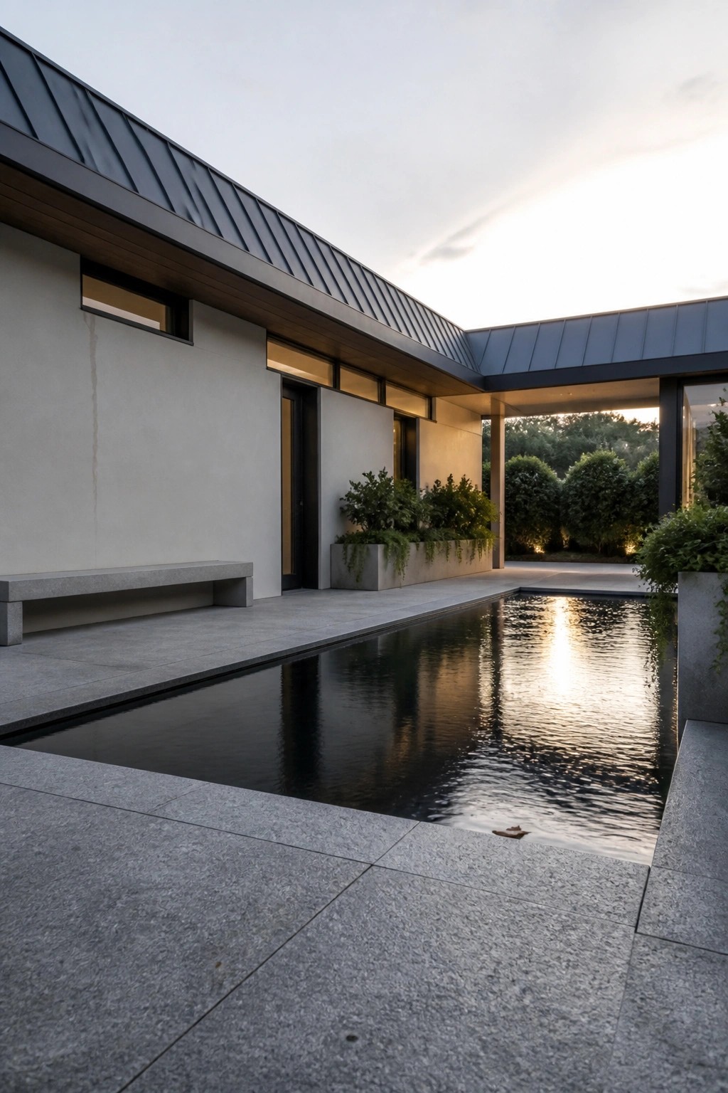 Modern house exterior with a long overhanging dark metal roof, light stucco walls, and a reflecting pool along the concrete patio.