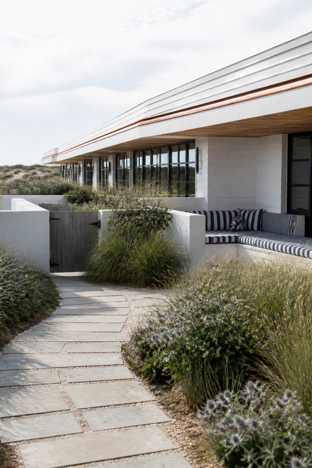 A modern single-story house with a long horizontal white roof edged in copper, black-framed windows, and a stone path leading past built-in outdoor seating with striped cushions.