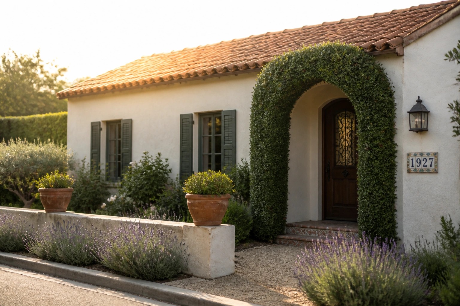 A small stucco house with a terracotta roof features an arched entry covered in green vines, a wooden door with iron details, two potted plants on pillars, and a gravel path leading to the entrance.