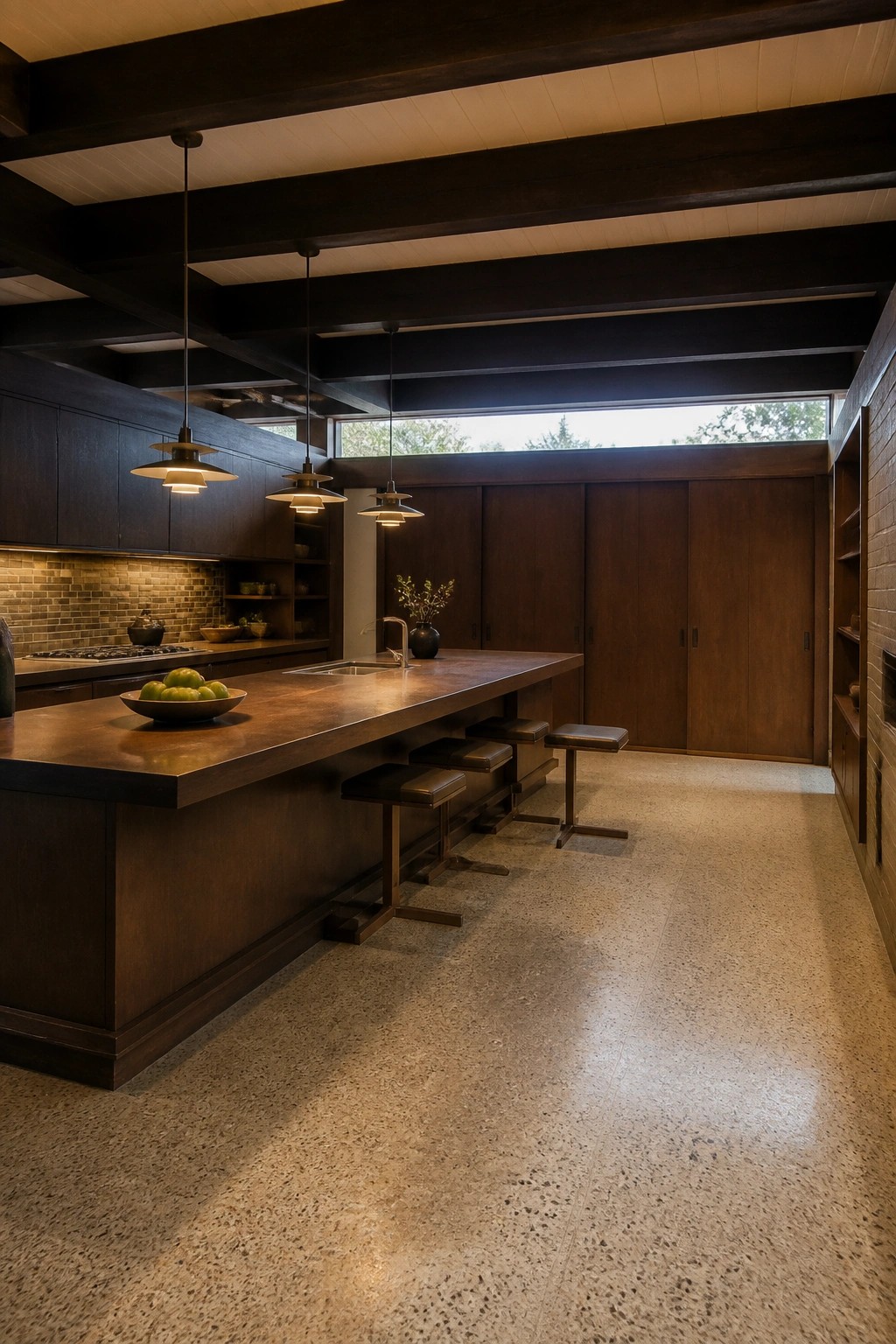 A kitchen interior with a long wooden island, four low stools tucked underneath, dark built-in cabinets, three pendant lights, and a speckled terrazzo floor.