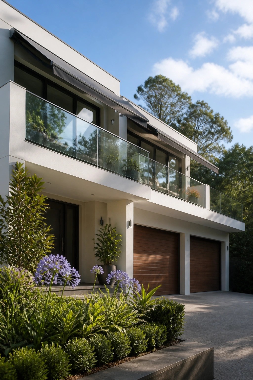 A modern two-story house with white walls, glass balconies, dark awnings, and two wooden garage doors, with landscaping in the foreground.