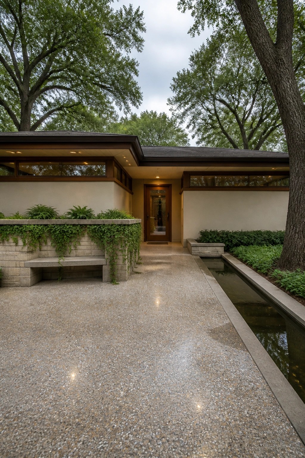 A long textured concrete path leads to a wooden door on a modern house exterior, with a narrow water channel on the right and stone planters with hanging vines on the left.
