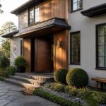A contemporary two-story house with vertical wood siding on the left side of the entry, a dark wooden door under a black metal canopy, large black-framed windows, and stone steps leading to a landscaped front yard.