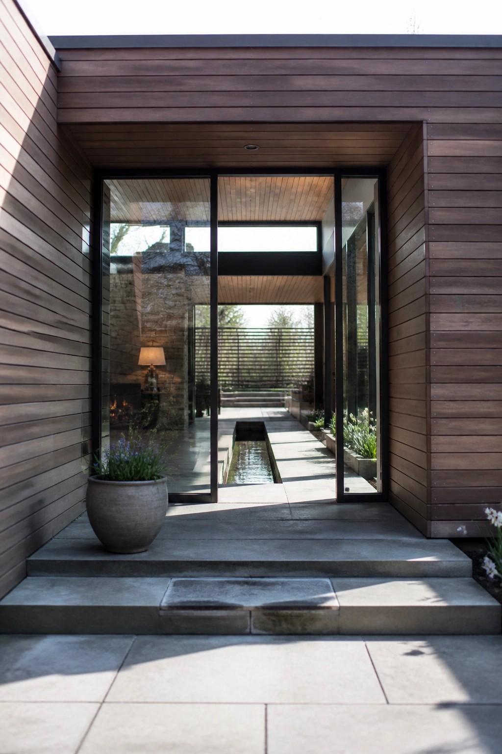 Front entrance of a modern house with large black-framed glass doors, wooden siding, concrete steps, and a view through to an interior stone fireplace and an outdoor water feature with garden beyond.