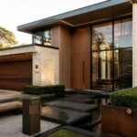 A contemporary house exterior with a wooden front door, vertical wood slats, light stone walls, large windows, and a paved entry path lined with plants and a metal planter.