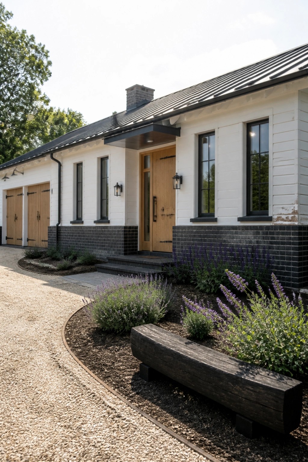A white farmhouse with black brick foundation, wooden front door and garage doors, black metal roof, and lavender plants along the front.
