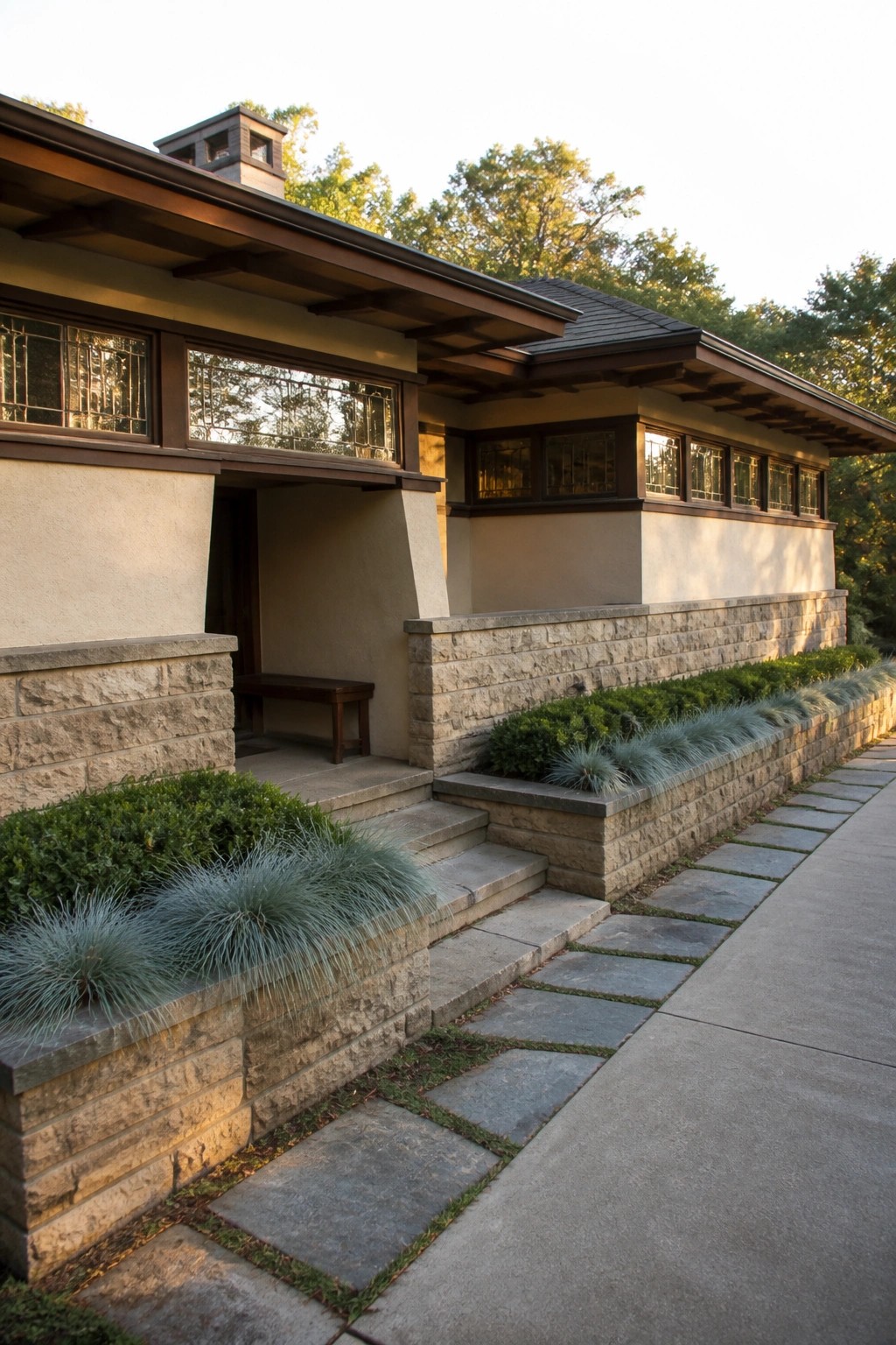A prairie style house exterior featuring horizontal windows, dark wood trim, stucco walls, and layered stone retaining walls with shrubs and ornamental grasses beside a stone pathway.
