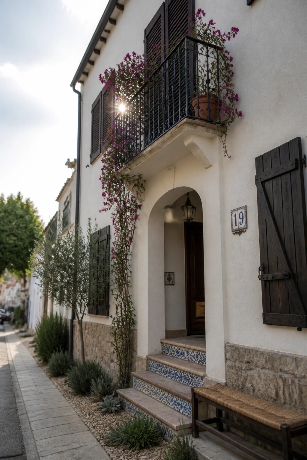 White stucco house exterior with dark shutters, a black iron balcony draped in purple flowering vines, an arched doorway with blue and white tiled steps, and a wooden bench beside the entrance.