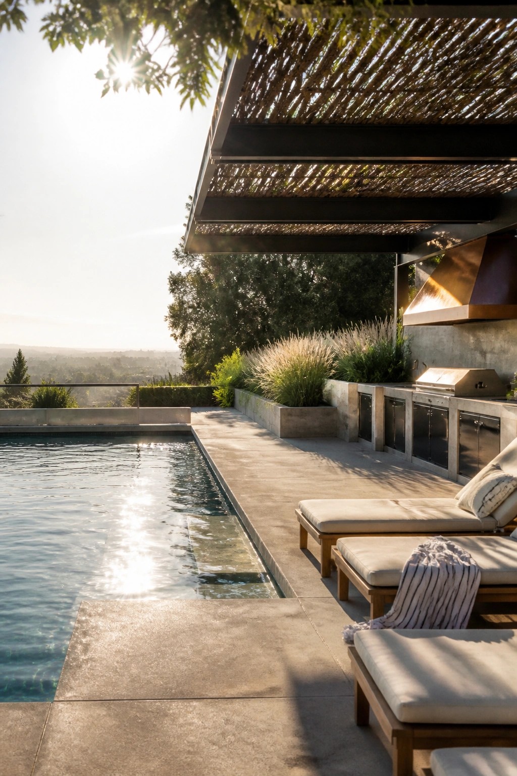 An outdoor poolside patio with lounge chairs, an outdoor kitchen under a large woven overhang, and distant landscape views.