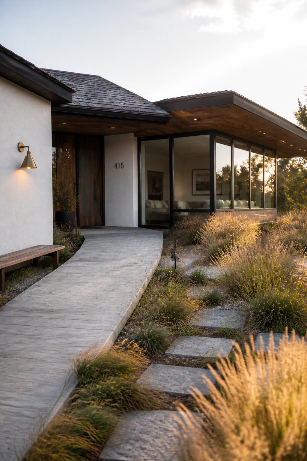 Modern ranch house exterior with white walls, dark wood trim, a pitched roof on the left, and a flat roof extension with large glass windows on the right, approached by a curved concrete path and stone steps through dry grasses.