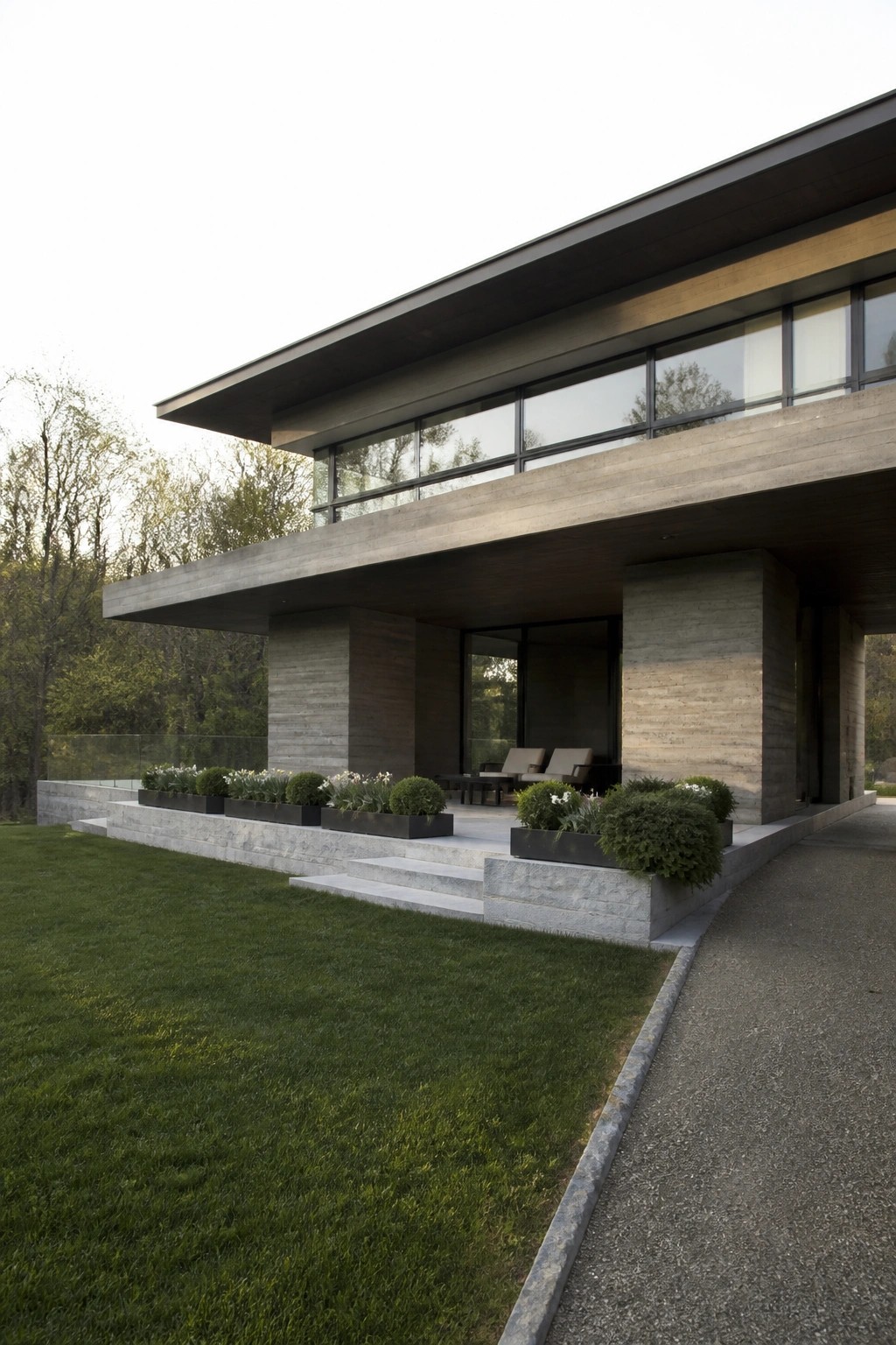Modern concrete house with large windows, patio seating, and green lawn