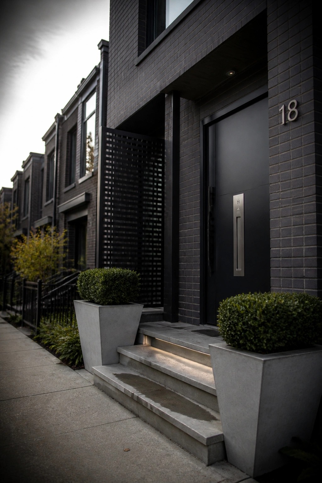 Modern dark brick townhouse with black door 18, lit steps, and gray planters.