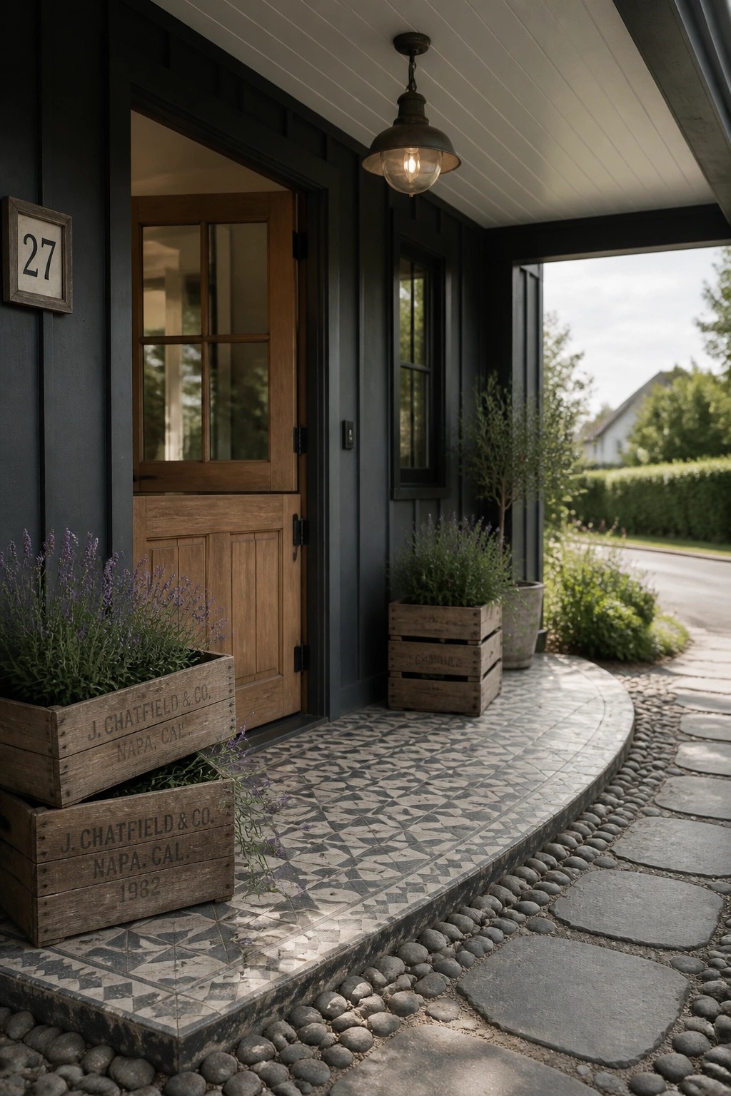A dark exterior entry with a wooden door, patterned tile porch, stacked wooden crates filled with lavender, and a stone pathway.