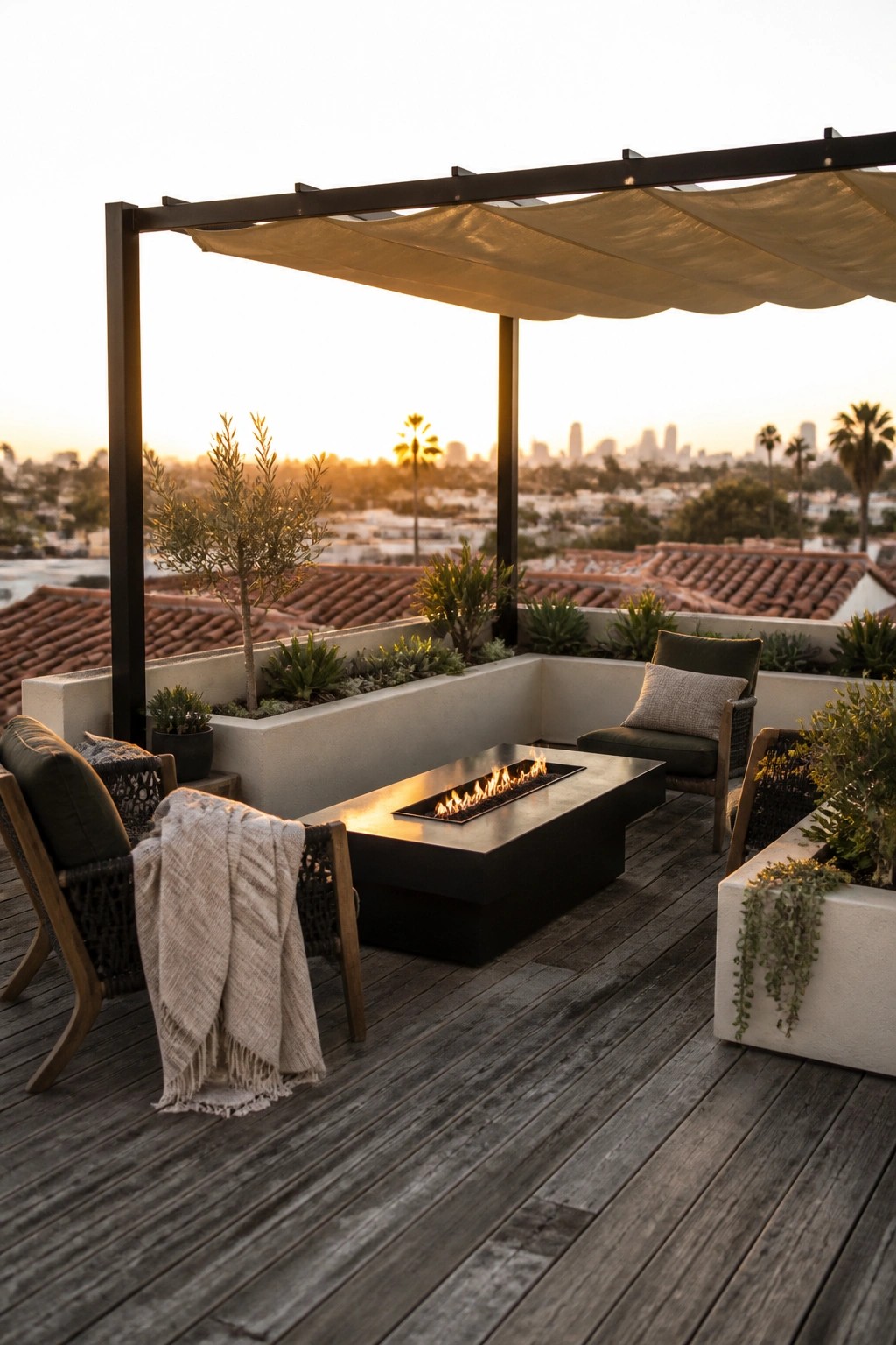 A rooftop terrace featuring a black metal pergola with beige fabric shade, a black rectangular fire pit on gray wood decking, woven chairs with cushions, and potted plants overlooking rooftops and a city skyline at sunset.
