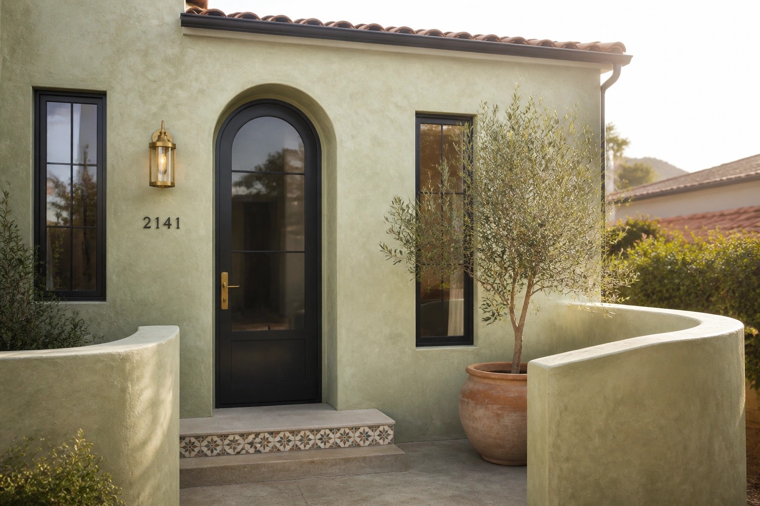 A light stucco house exterior with a black arched door, wall lantern, potted olive tree, and tiled step riser.